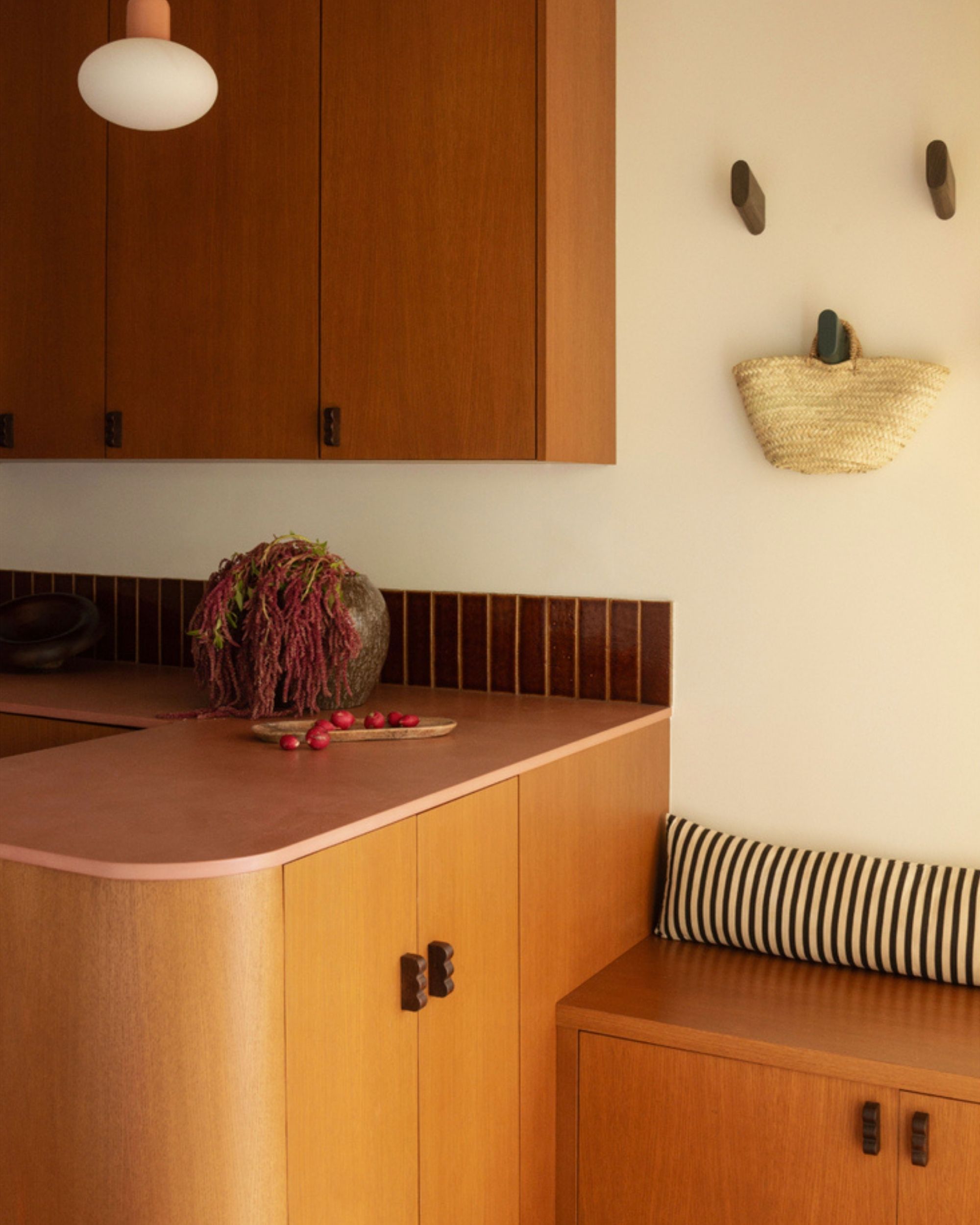 The corner counter of a kitchen that has orange wood cabinets and a pink countertop. There is a bench seat with a striped pillow and hooks on the wall with a straw bag hanging form one.
