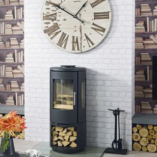 Woodburner in front of a fake feature wall showing white brick in the middle and a rustic look brown clock with roman numerals, and bookshelves either side