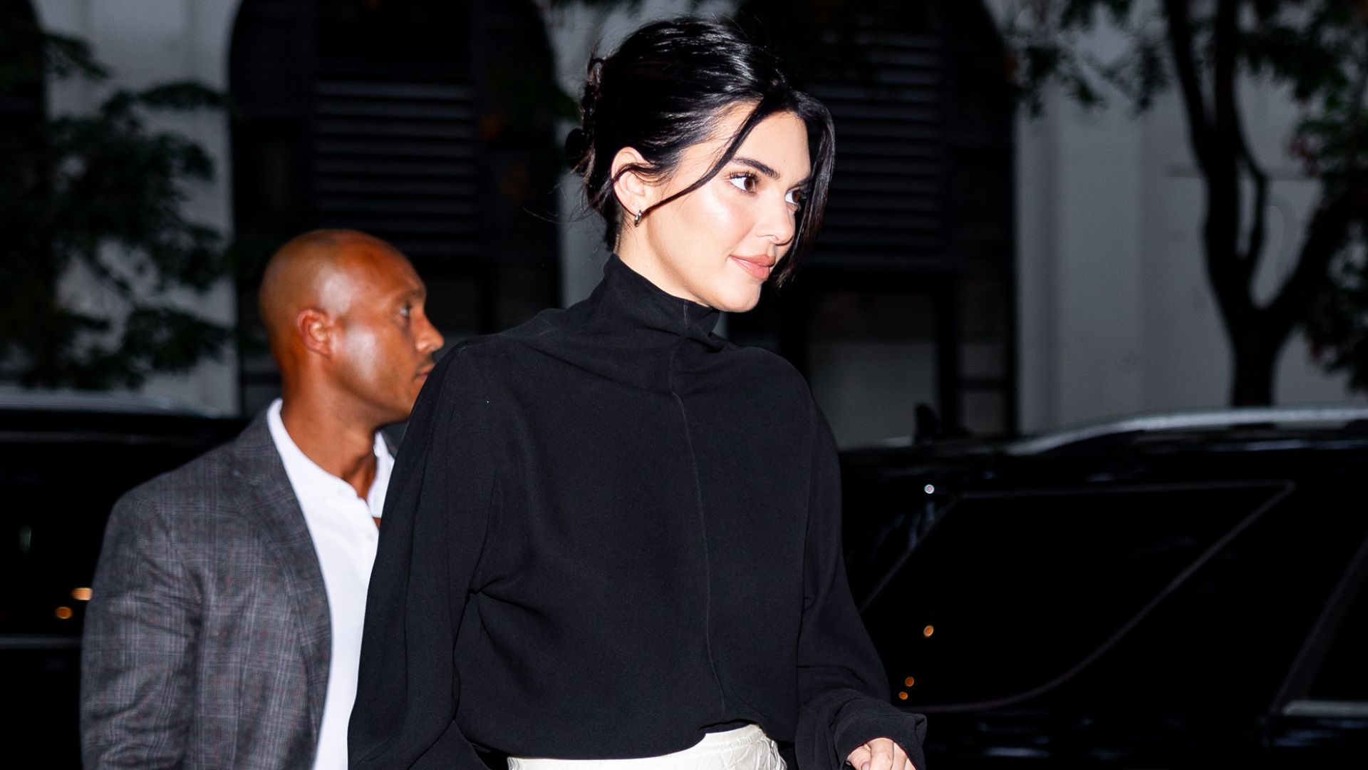 Kendall Jenner walks outside wearing a black knit, black midi skirt and mid-heel pumps.