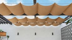 A beige retractable sun shade on a black pergola in front of a white wall with a black privacy fence to the side and a blue sky above 