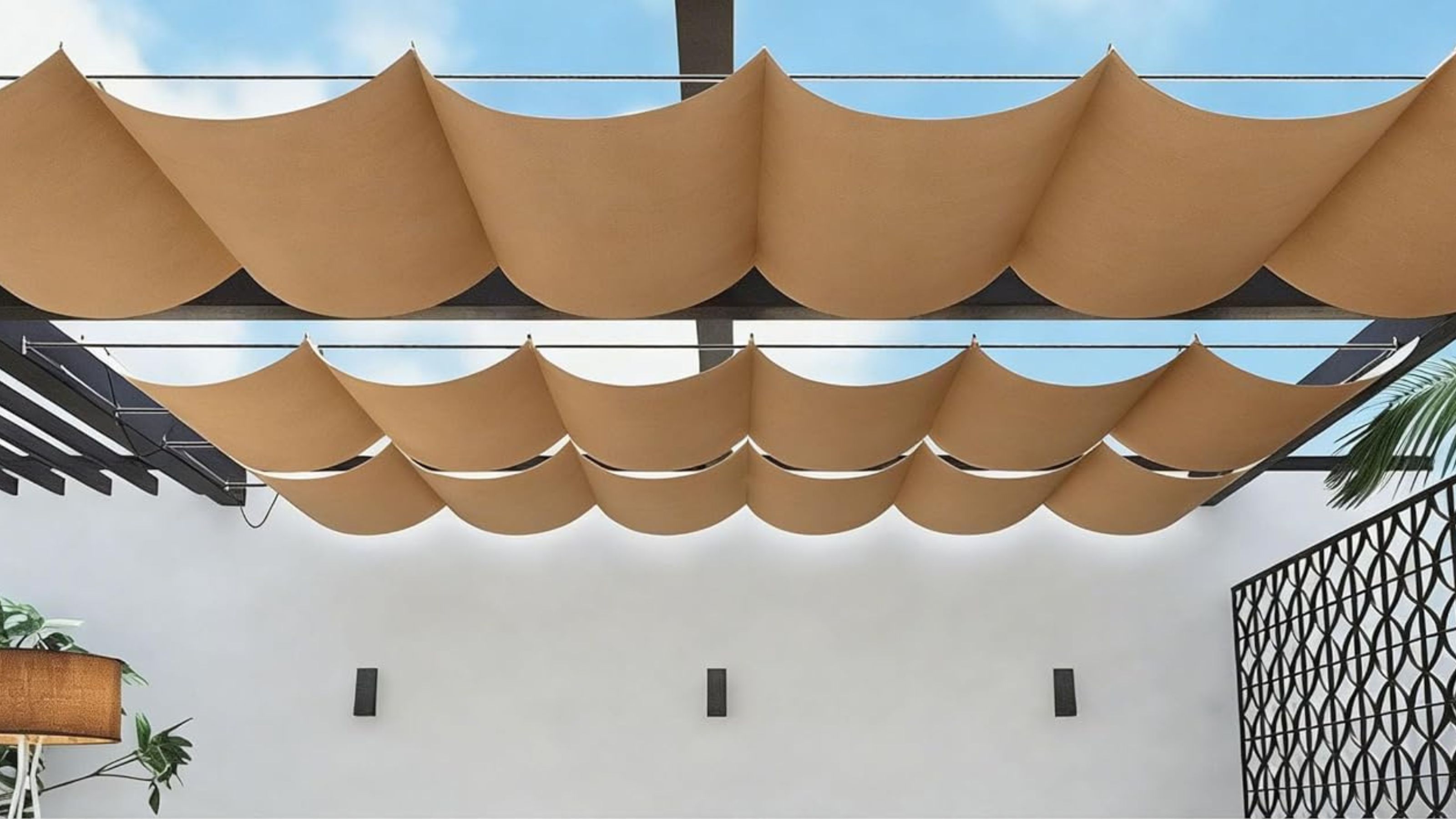 A beige retractable sun shade on a black pergola in front of a white wall with a black privacy fence to the side and a blue sky above 