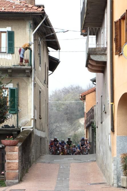 An elderly woman watches from above whilst the bunch passes through a narrow village entrance