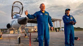 two astronauts in flight suits standing in front of a t-38 jet with cockpit open. they are on a tarmac with a structure in behind and buildings far in the back