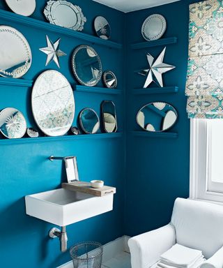 Blue painted bathroom with selection of ornate mirrors on shelving, rectangular sink, wooden board, white antique armchair, towels, wire waste basket, white and blue patterned blind