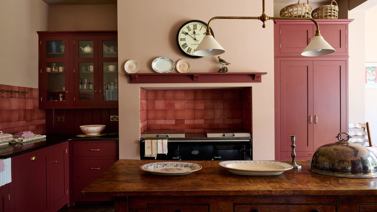 A pink kitchen with marble kitchen island, glass cabinets, a black AGA and large clock above