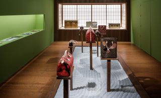 Louis Vuitton’s Olivier Saillard-curated exhibition, ‘Volez, Voguez, Voyagez – Louis Vuitton’. Various sized and coloured handbags displayed on wooden square poles in a room with green wall shelves and cupboards and a luggage display inset into a wooden wall.