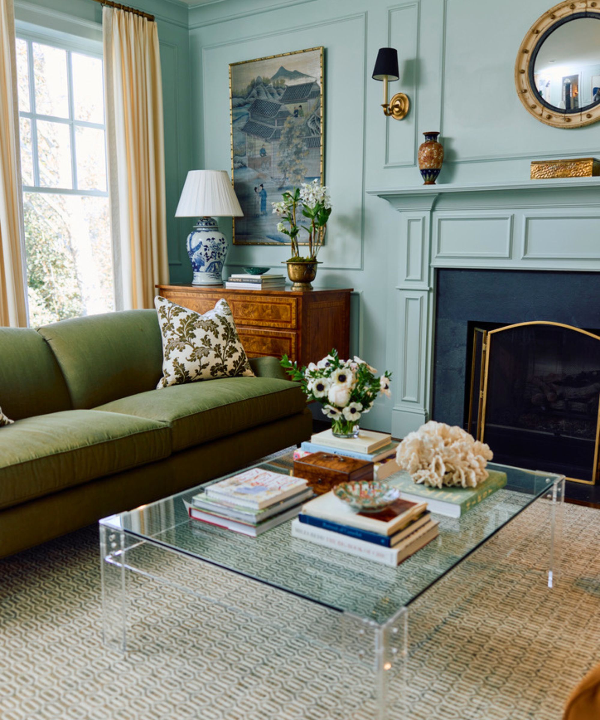 Living room with pale blue walls, grand fireplace, acrylic coffee table, green sofa, mahogany dresser and ivory drapes