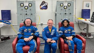three astronauts in a row, with spacesuits. two of them are sitting down on chairs. one in the middle is kneeling. behind them are panels and a flight patch for crew flight test showing a rocket