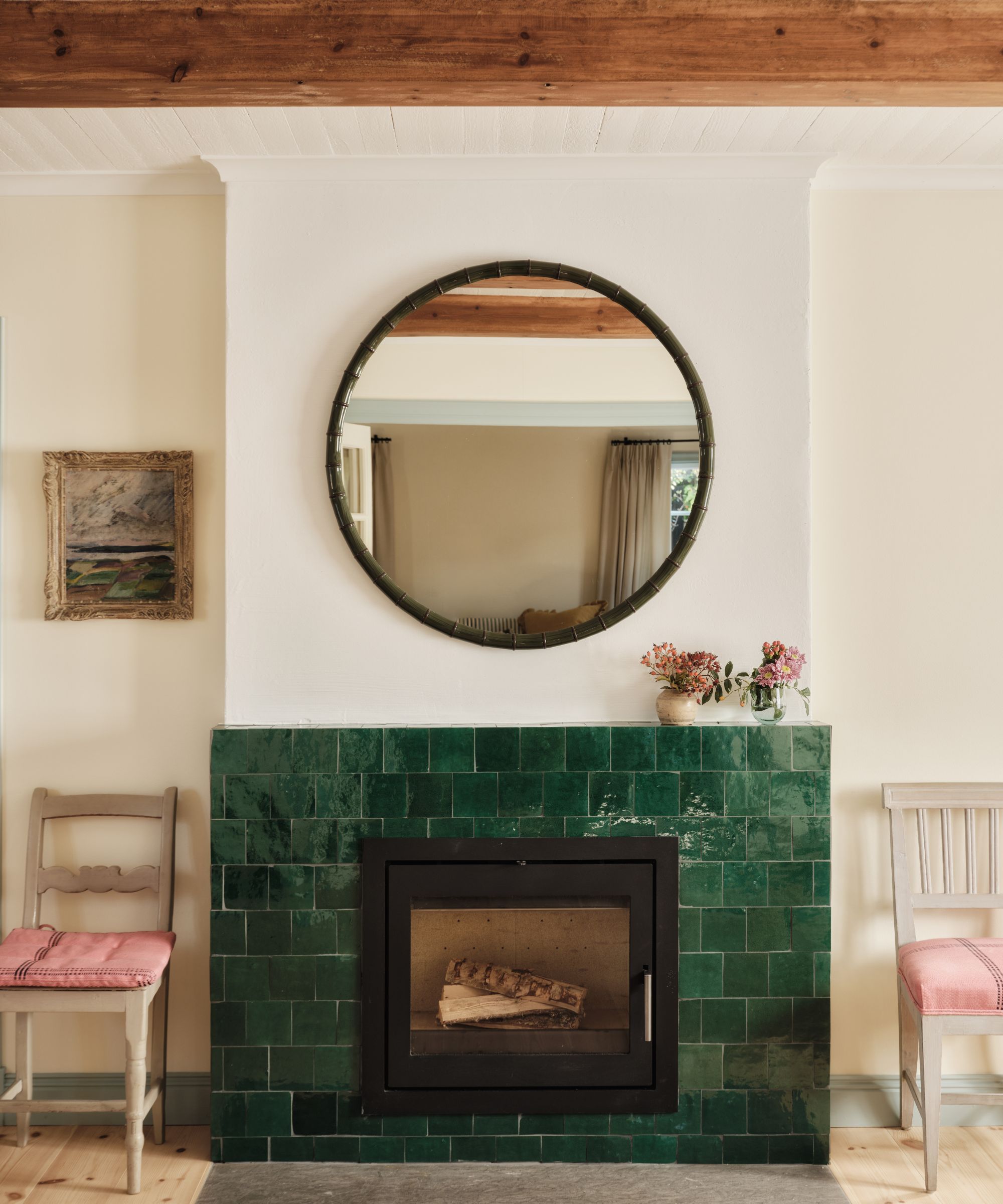White living room with a fireplace surrounded by glossy green tiles and a circular mirror hanging above. Two chairs sit in the alcoves with pink cushions