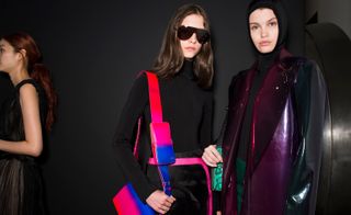 Models wear PVC coats and black turtlenecks. One model holds an ombre pink and blue leather bag