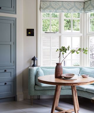 Corner of a blue kitchen with bay window, upholstered blue bench, round wooden dining table, blue patterned blind