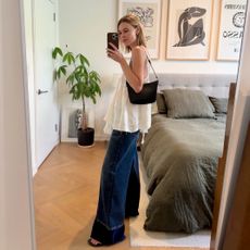 Stylist and fashion editor Natalie Cantell taking a mirror selfie in New York wearing Stella McCartney denim, a white flowy summer top, and a black Celine Croque shoulder bag, and brown flip-flops.