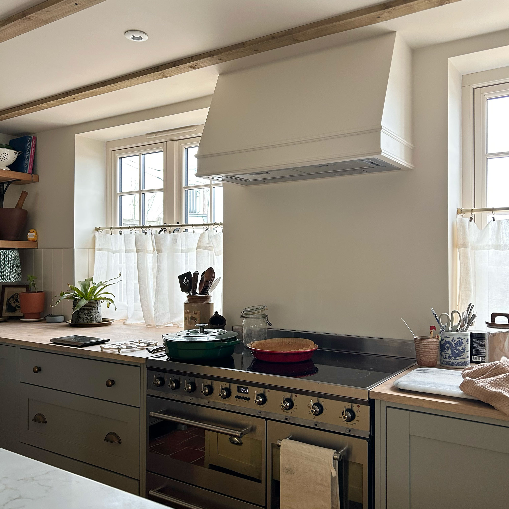 Oven in kitchen topped with white cooker hood, framed with cafe-curtained windows on either side