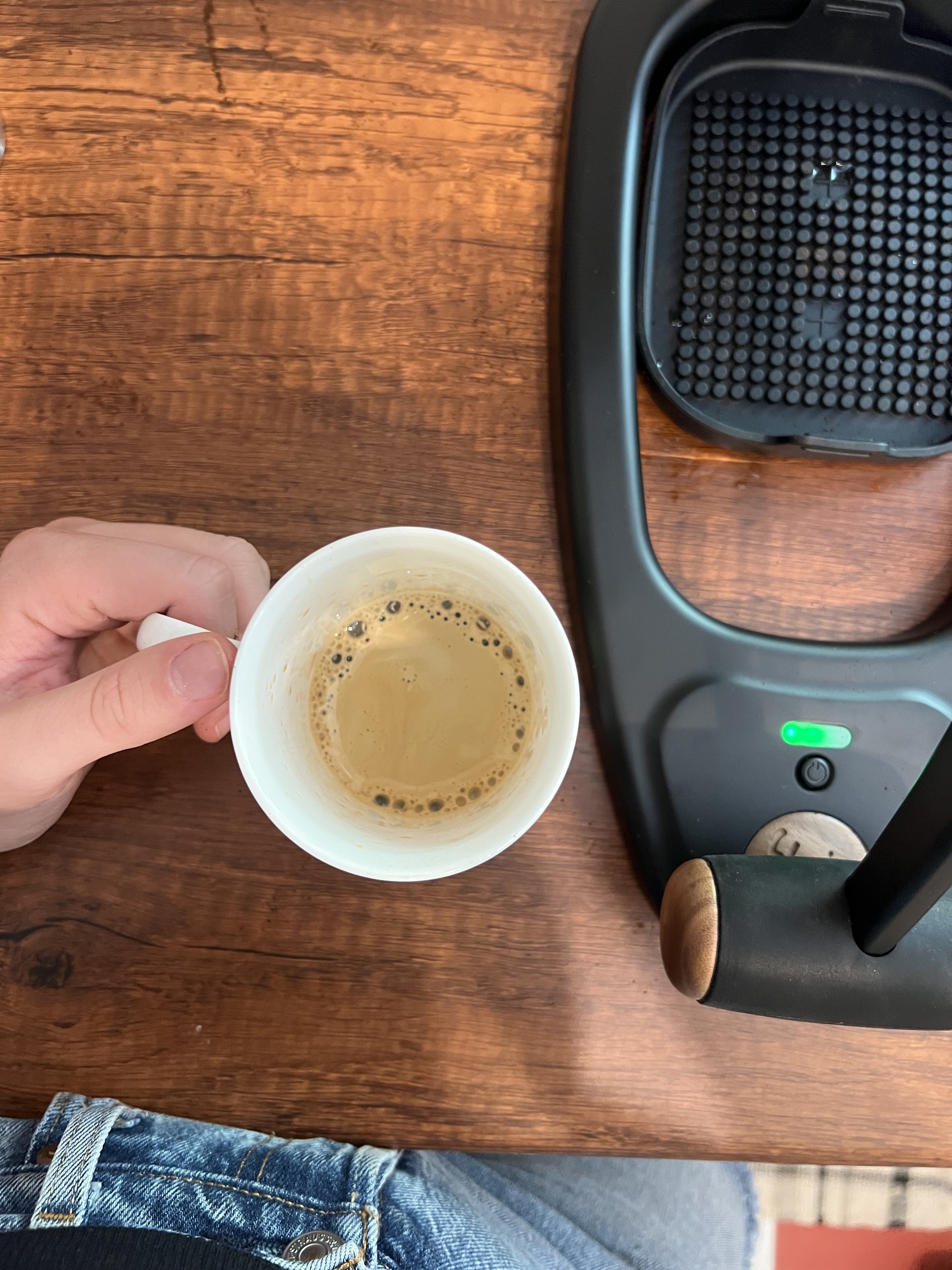A hand holding a white coffee mug with an espresso shot in it next to a black manual coffee machine on a wooden countertop.