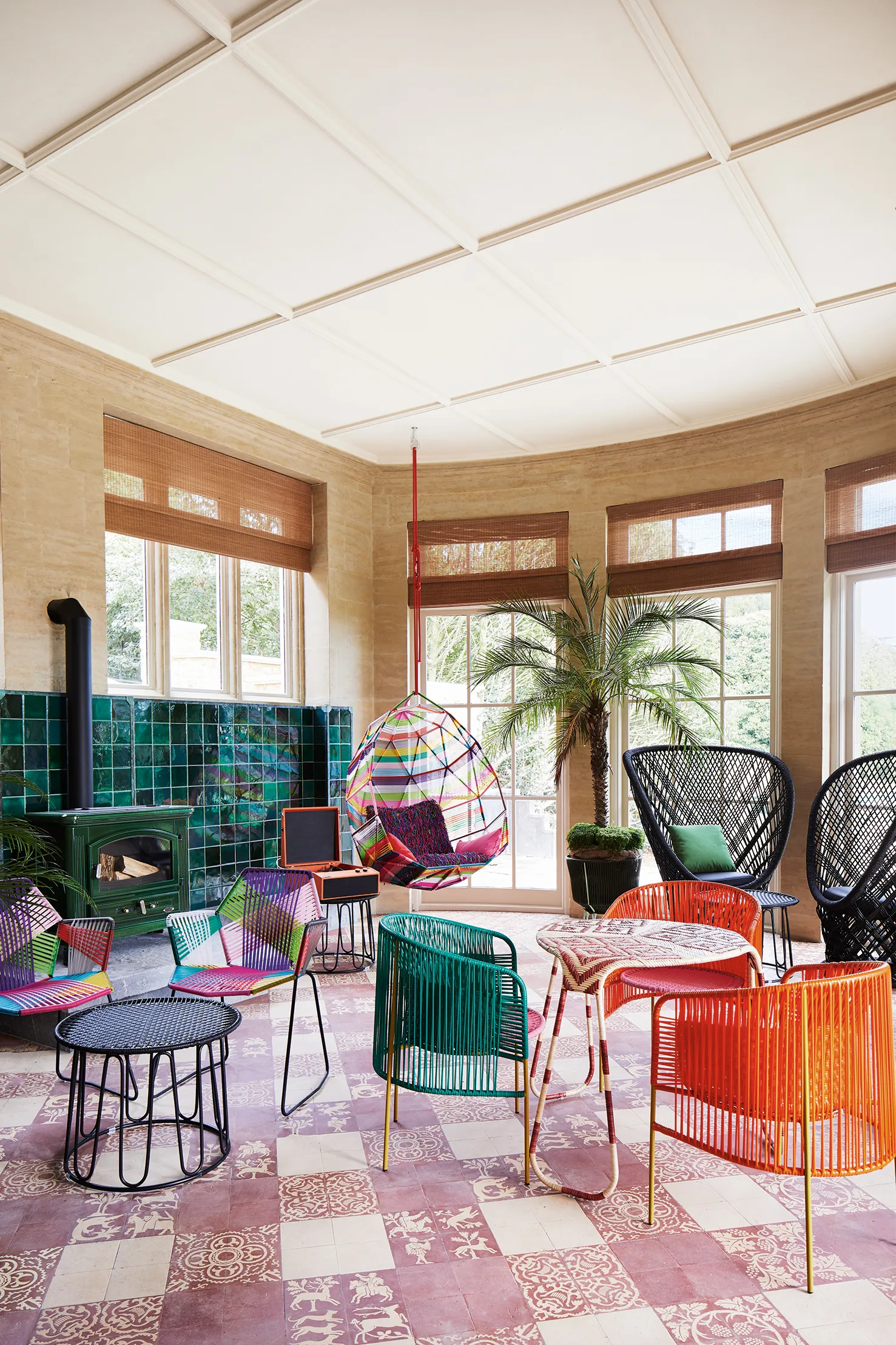 A lively furnished hotel lobby features eye-catching sculptural pieces in painted rattan that clash with the petrol green wall tiling, patterned floors, and wooden walls of the farmhouse it is housed in.