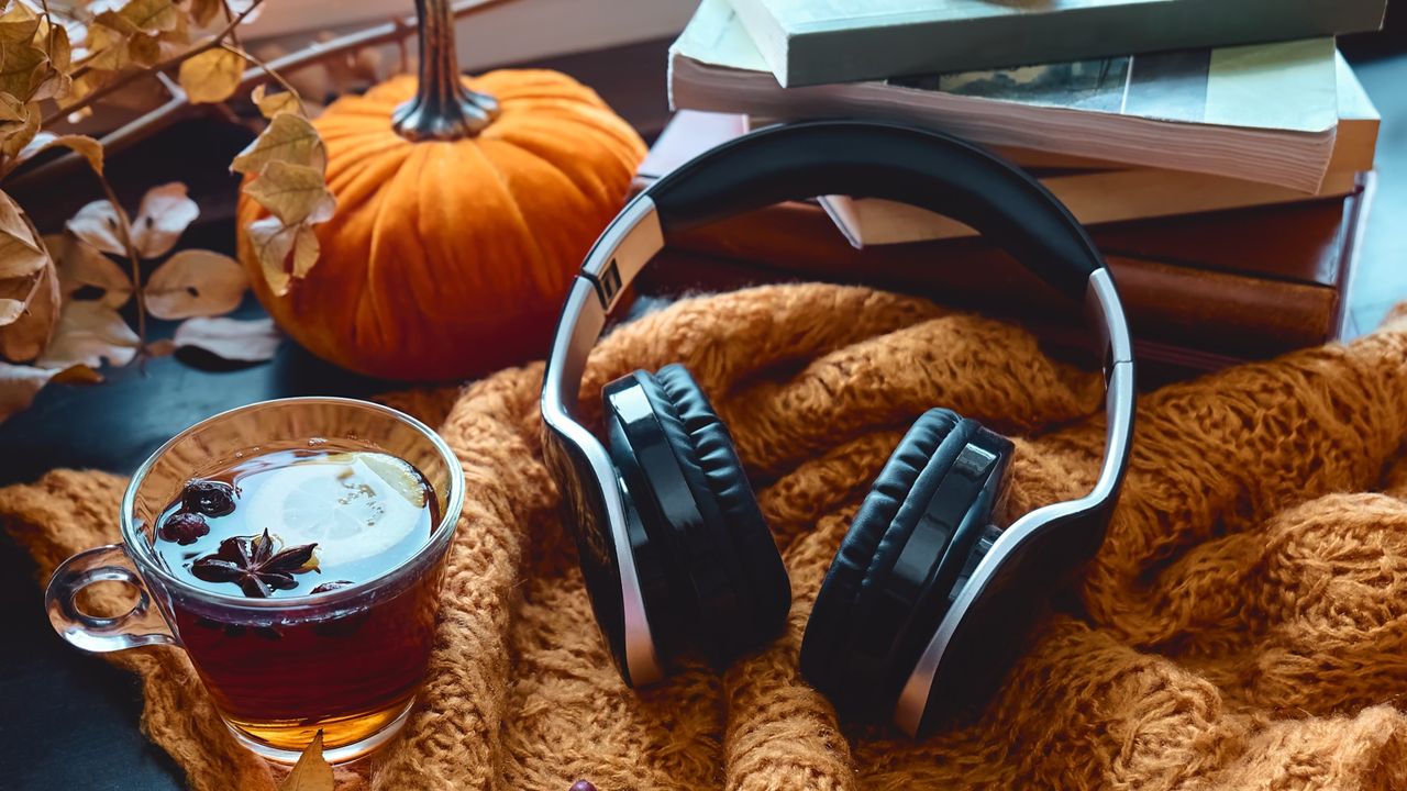 A pair of headphones resting near a fall-themed cup of tea and a pumpkin.