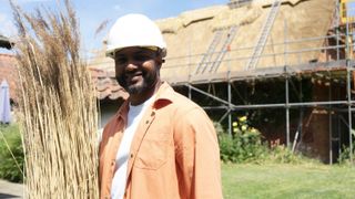 JB Gill in a white top and peach shirt and white hard hat holding some thatch outside a thatched house in Summer on the Farm.