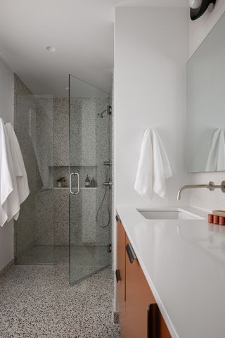 Image of a white, modern bathroom with burnt orange cabinetry and white countertops. The floors bleed into the shower tiles which are all a brown and black terrazzo material