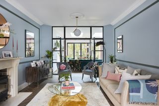 Light blue living room with dark parquet flooring, cream sofa and rug, glass coffee table and internal glazing into kitchen-diner