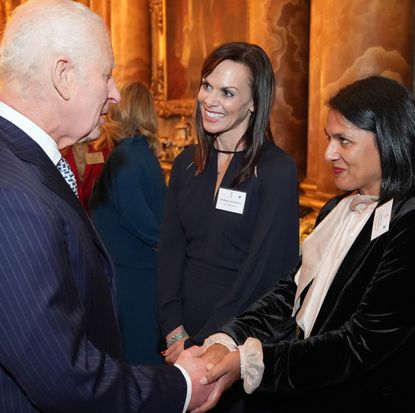 King Charles wearing a blue pinstriped suit shaking hands with Naveed Chowdhary-Flatt who is wearing a black velvet blazer and cream pussybow blouse