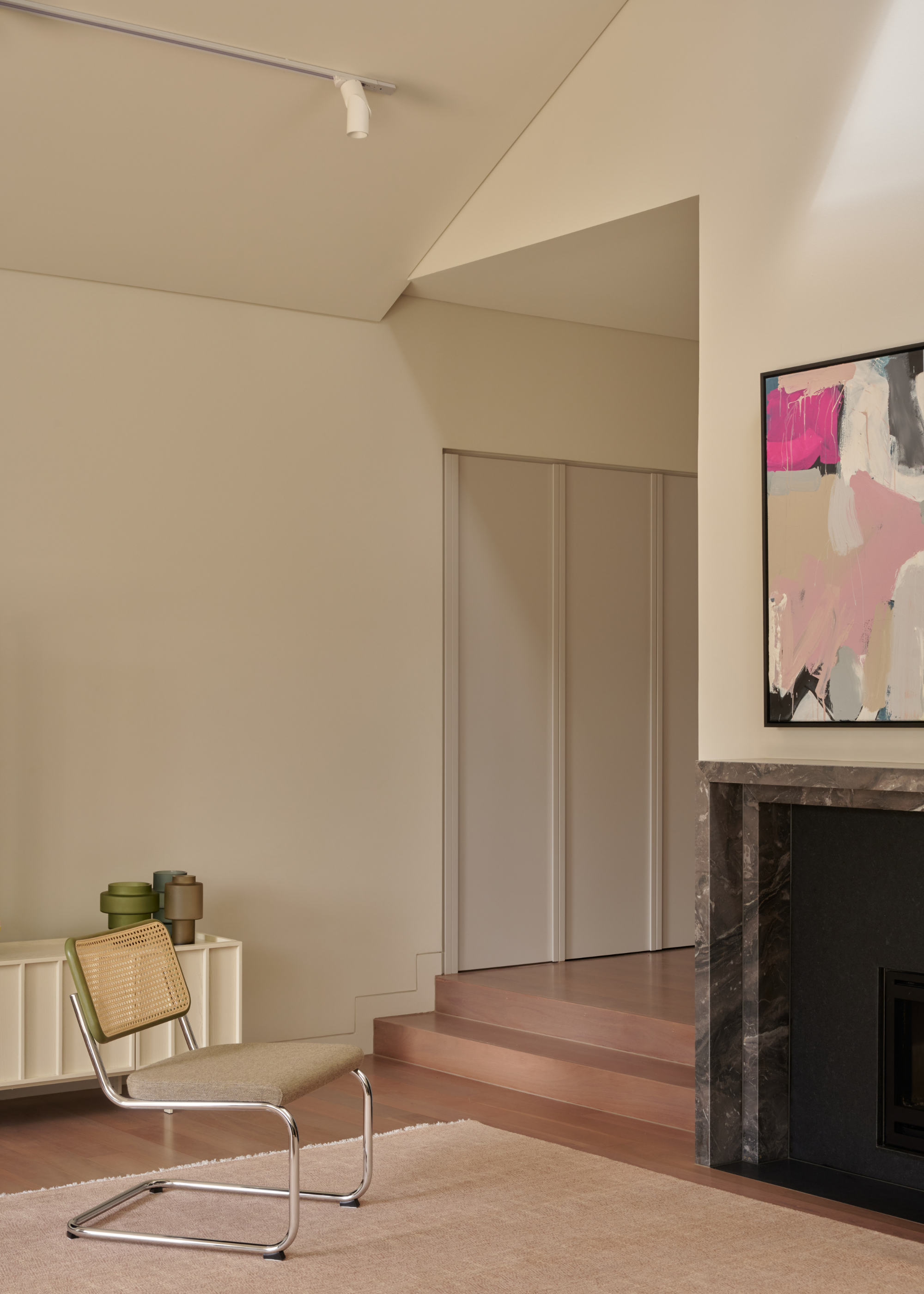 A white living room with a white sideboard featuring colored vases, a metal chair with a rattan back rest, a framed colorful painting over a stone fireplace