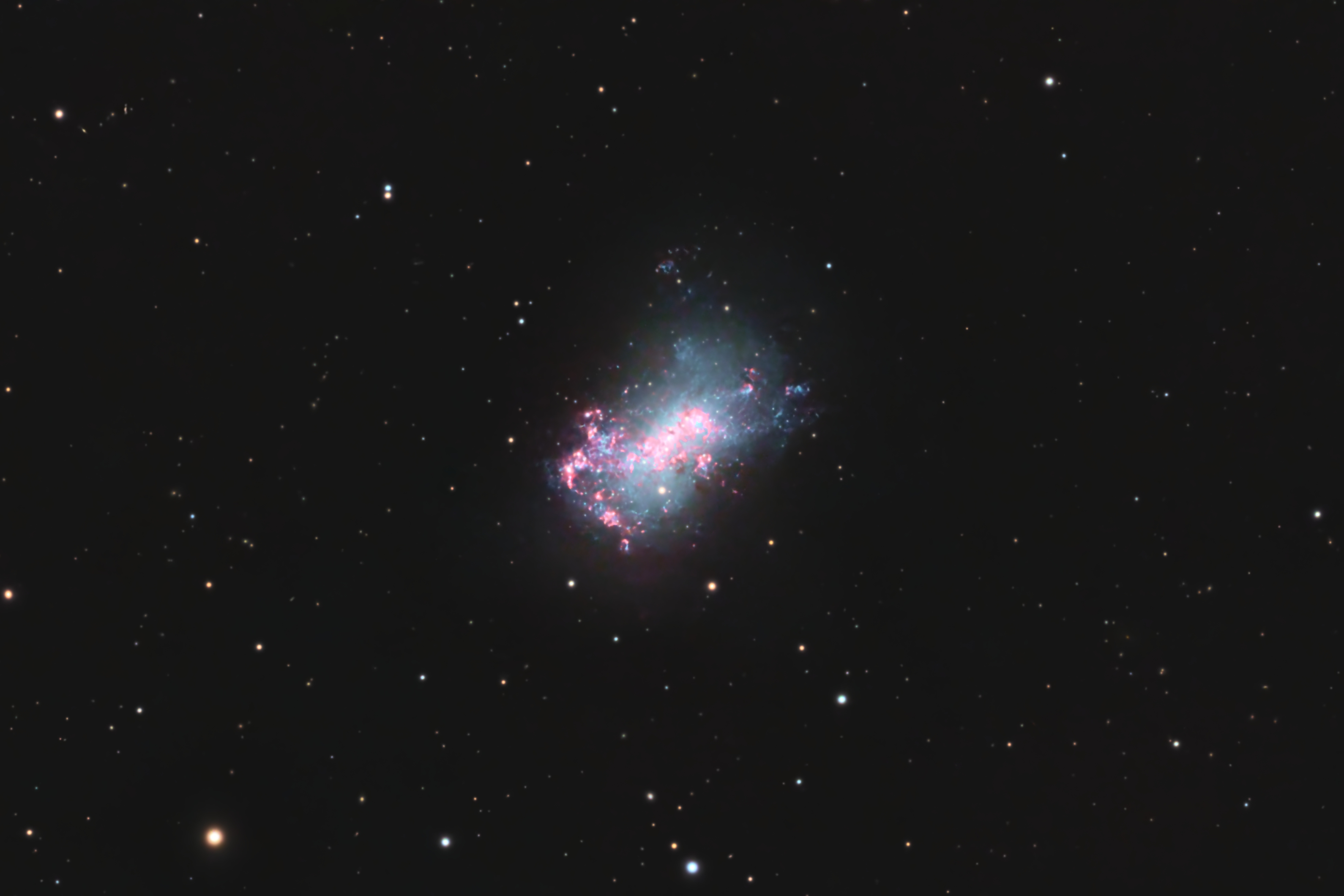 distant pink hued clouds are asymmetrically dispersed around a light blue cloudy haze in the blackness of space. The dwarf galaxy NGC 4449 is the first dwarf galaxy with an identified stellar stream (faintly seen at the lower right, and in inset). The star stream represents the remains of a smaller satellite galaxy merging with NGC 4449.