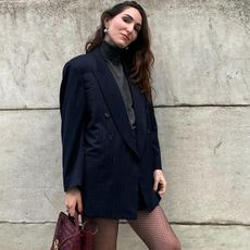 fashion editor wearing black blazer, black mini skirt, and grey turtleneck against brick wall