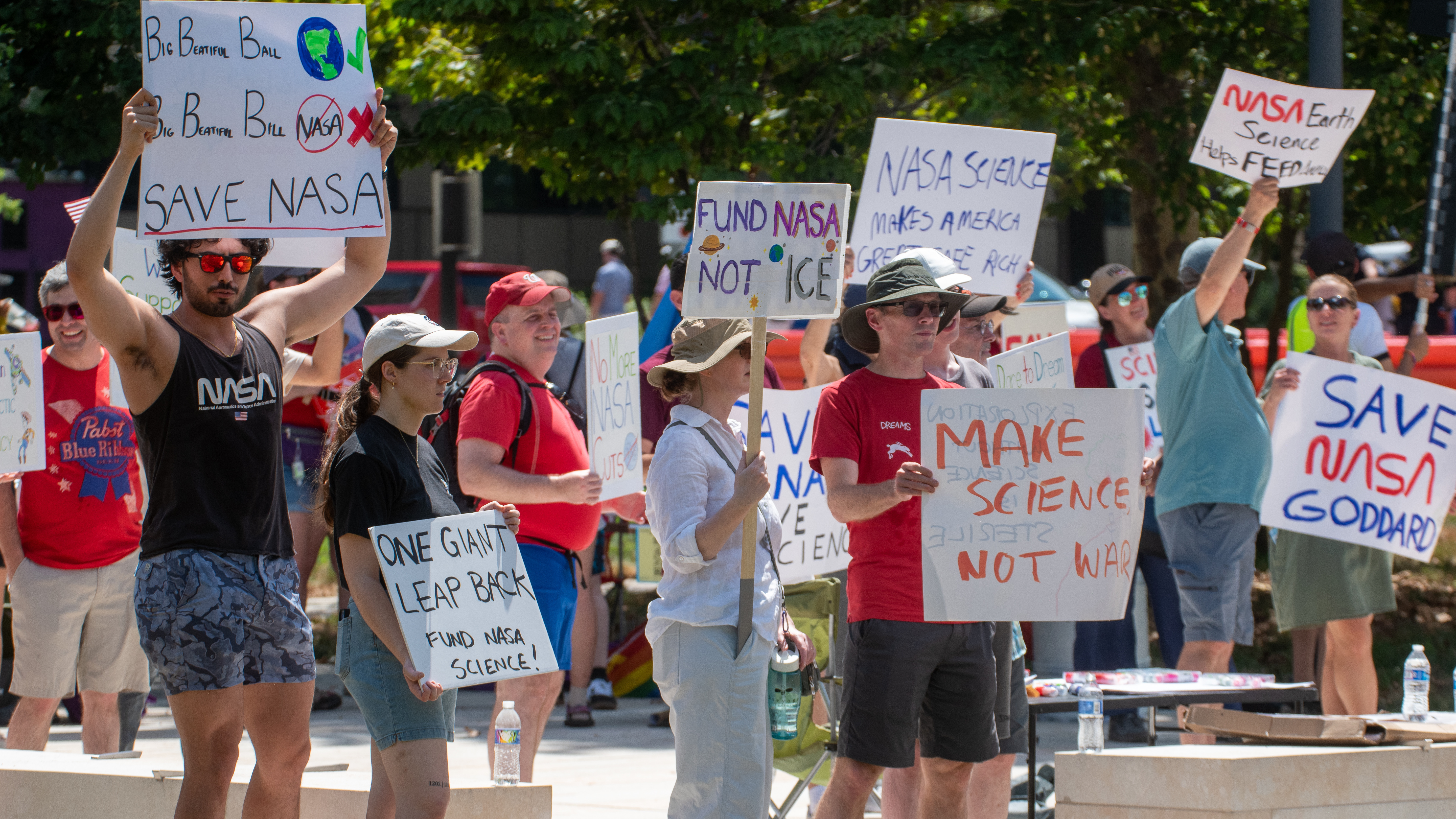 People hold signs outside on a sunny day in protest of NASA budget cuts, July 20, 2025.