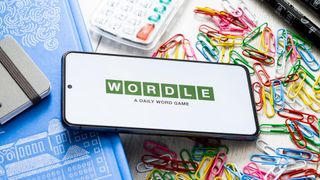 A phone displaying the Wordle logo sitting on a table surrounded by paperclips, pens and notebooks
