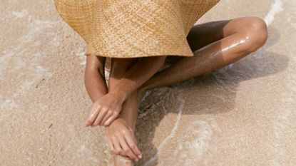 Woman in a straw hat sat on the beach with the sea around her, shot from above