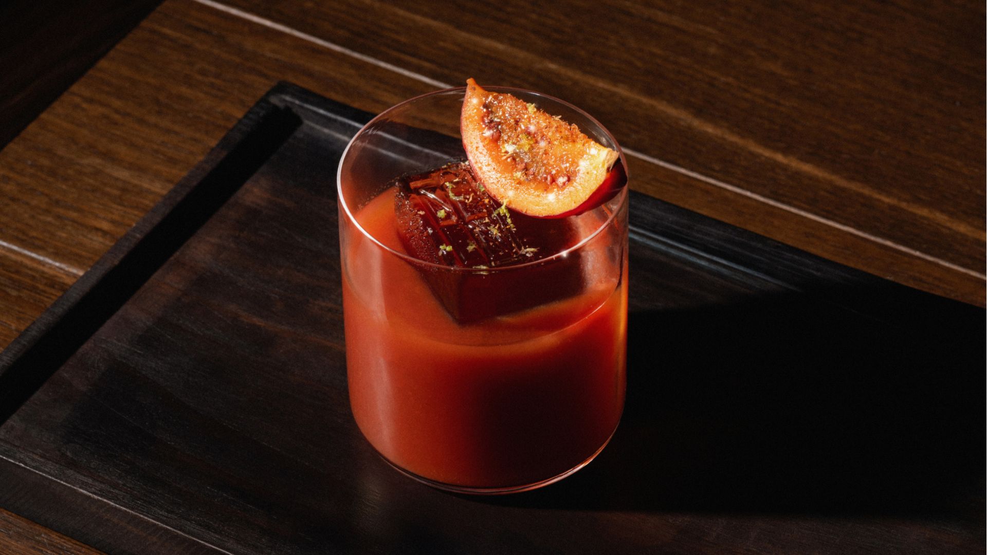 an image of a whisky cocktail on a brown wooden tray. The cocktail is a twist on a bloody mary, served in a glass tumbler with a large ice cube and a caramilsed peach slice garnish