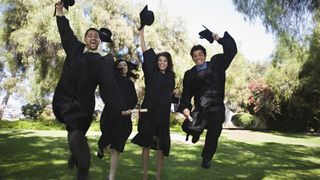 Graduates jumping and lifting hats, against a backdrop of green leafy trees.