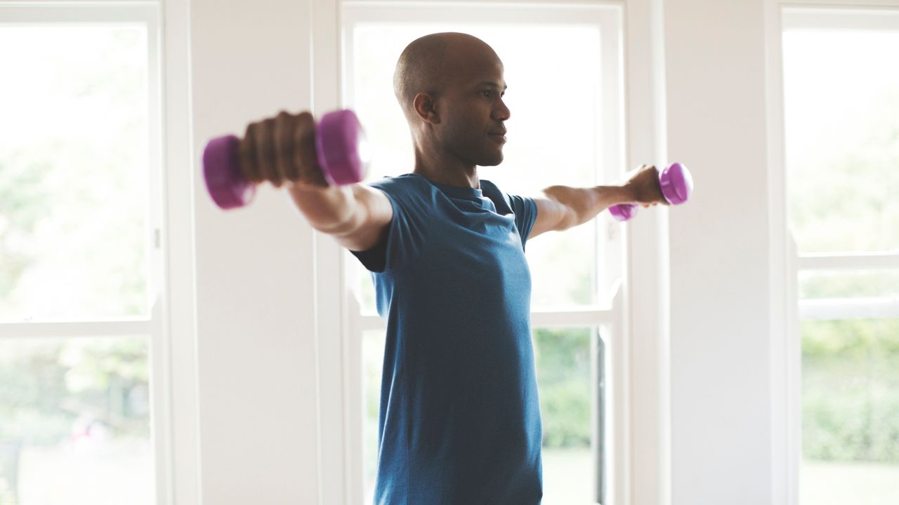 man facing sideways to the camera holding a light dumbbell in each hand held up laterally at shoulder-height. wearing a blue tshirt indoors with windows behind him. 