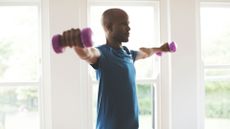 man facing sideways to the camera holding a light dumbbell in each hand held up laterally at shoulder-height. wearing a blue tshirt indoors with windows behind him. 