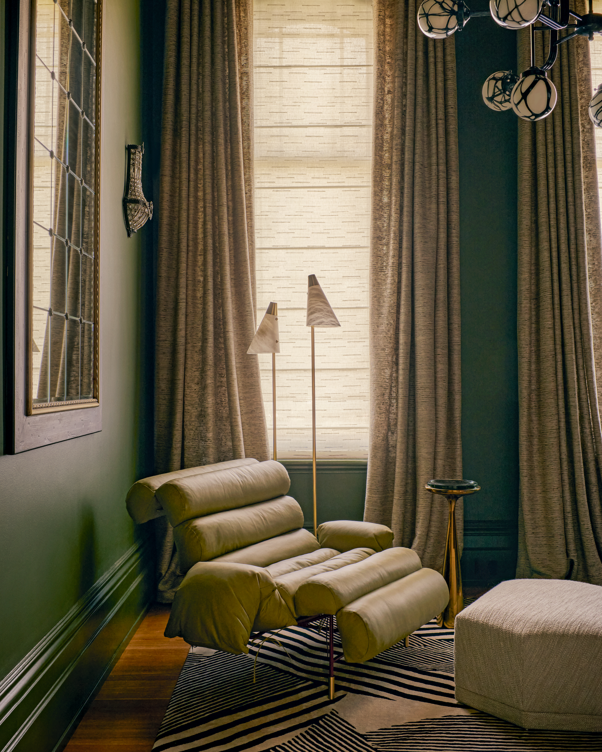 Library with mid green walls, green leather armchair, beige curtains and marble and brass floor lamps
