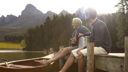 An older father relaxes with his adult son on a dock. Their bare feet are in the water or the gunnel of a canoe. They are drinking beers.