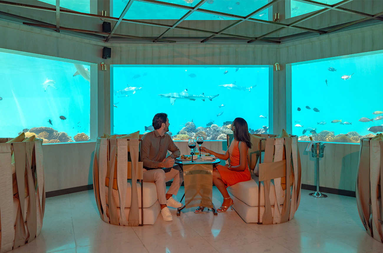 Two people sat at a table with food and glasses of wine, with a surrounding aquarium around them, blue water with lots of fish and coral swimming