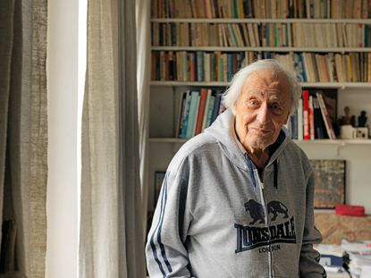 William Klein wearing a Lonsdale grey jumper standing in his home in front of a bookshelf