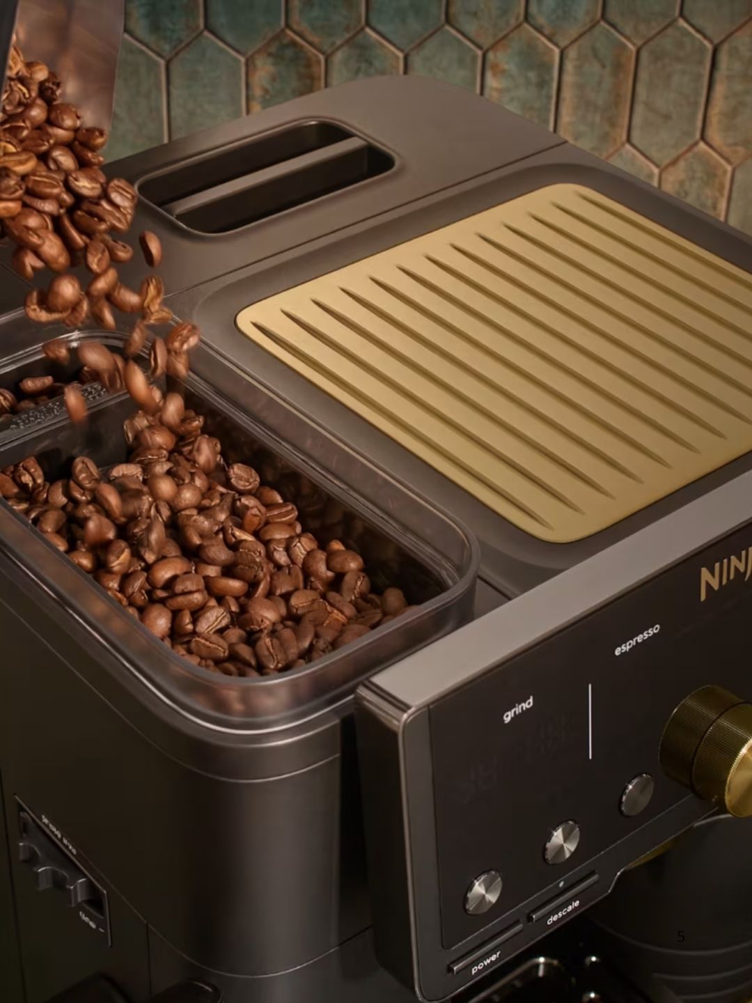 Close-up image of the top of a dark gray and gold espresso machine with coffee beans being poured into the hopper. 