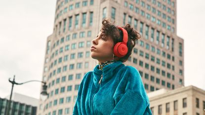 A woman sitting on a wall in a city, wearing a pair of the best headphones for travel – Beats