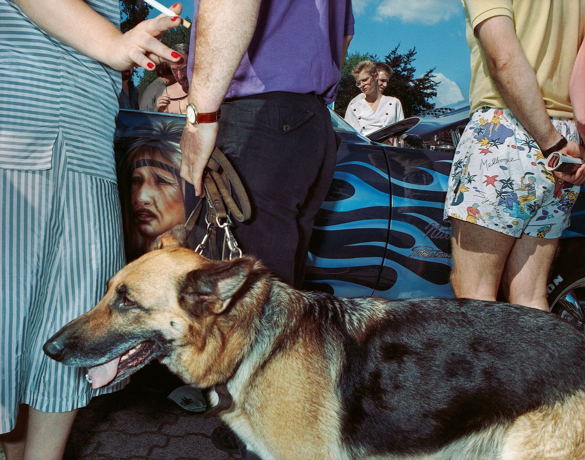 A German shepherd dog stands at the centre of a crowd in 1991 Cologne, surrounded by legs, shorts and a car airbrushed with blue flames and a woman's face.