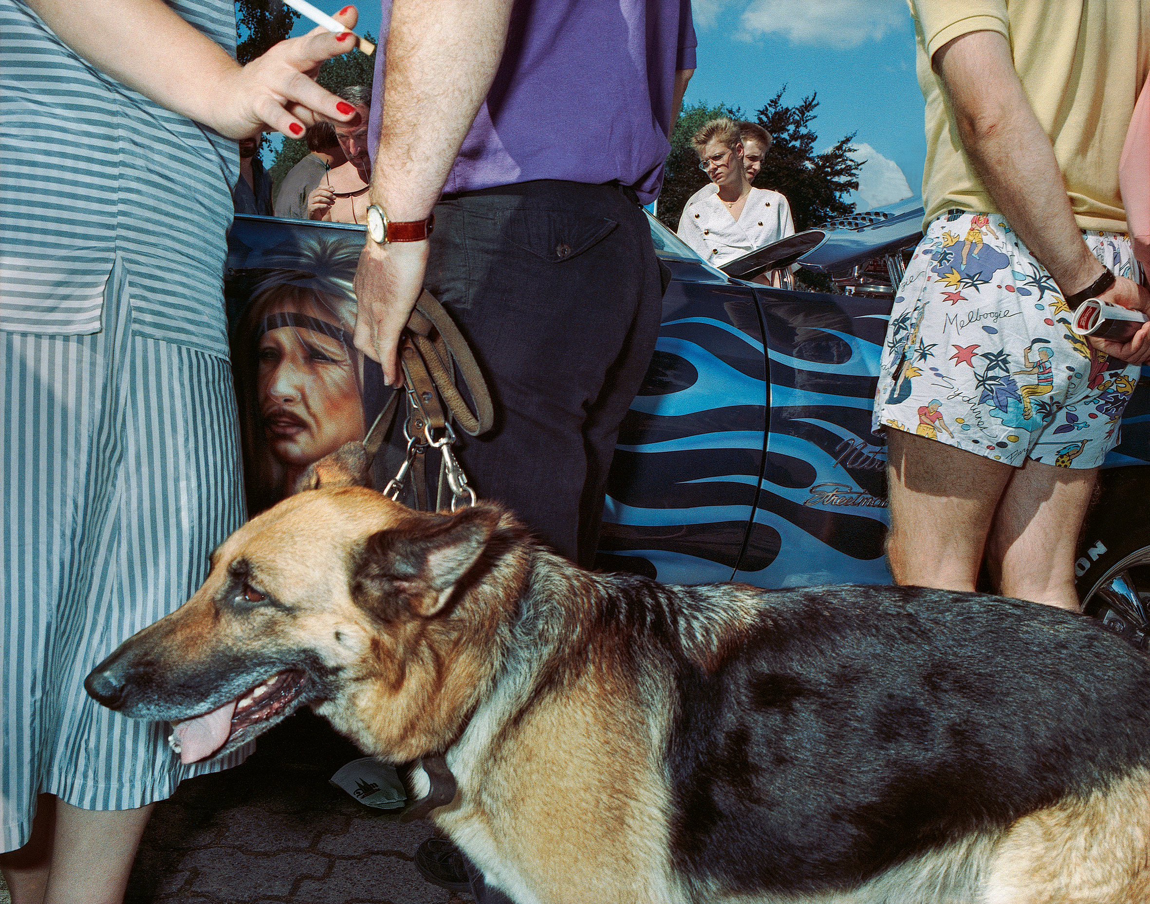 A German shepherd dog stands at the centre of a crowd in 1991 Cologne, surrounded by legs, shorts and a car airbrushed with blue flames and a woman's face.