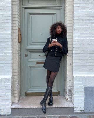 Woman wearing a black jacket, black mini skirt, black tights, and black calf-high boots standing in front of a doorway