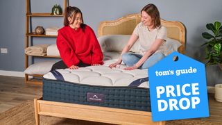 Two Tom's Guide sleep staff writers sit on the DreamCloud Luxury Hybrid mattress, with a blue price drop sales badge overlaid on the bottom right hand corner of the image