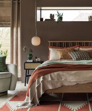 Cozy bedroom with brown painted walls, bed with cream, orange and blue linen, orange patterned rug, rattan and black wood bedside table, low hanging rounded glass pendant with striped design, boucle lounge chair