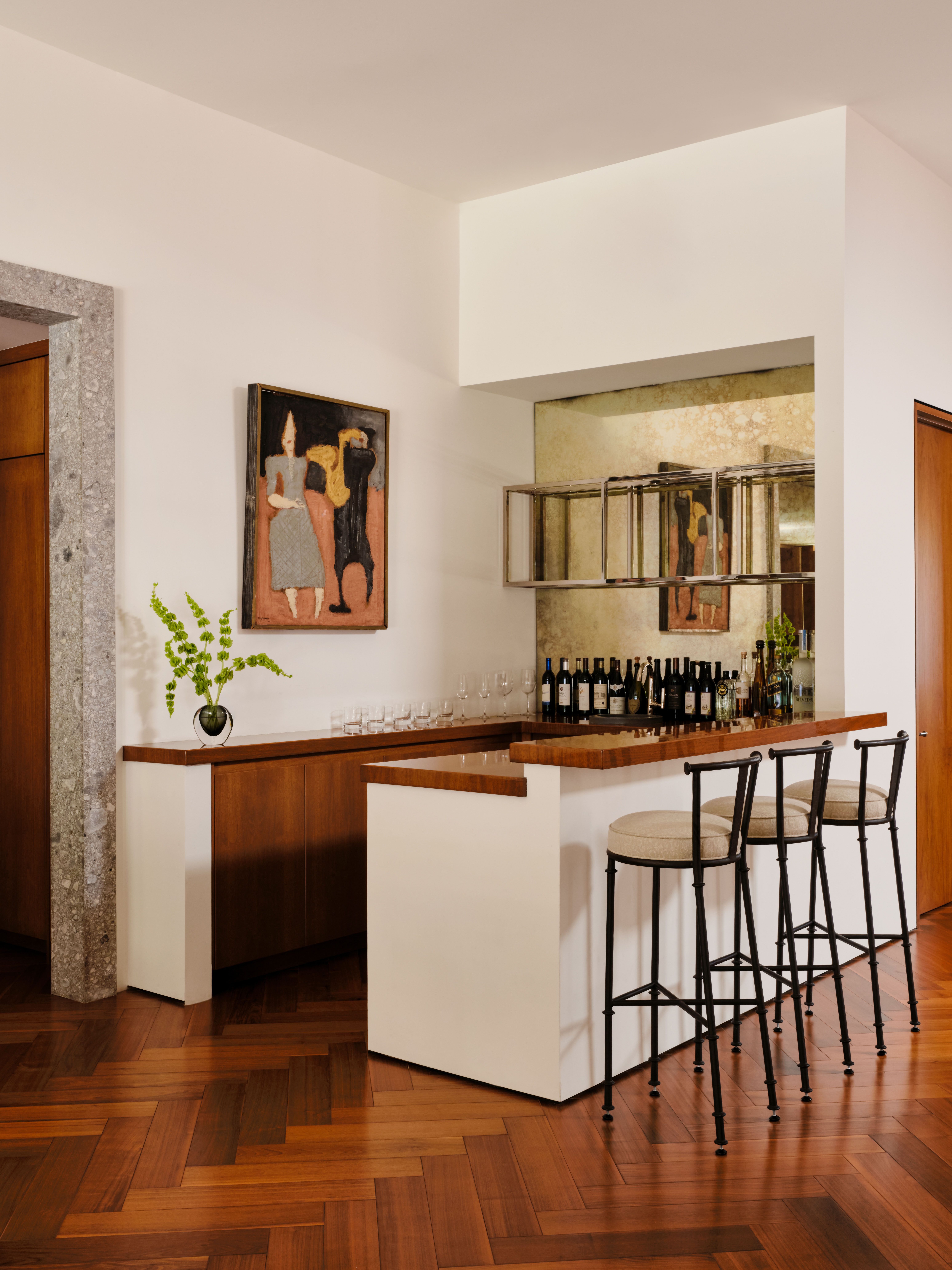 A house bar with tall wrought iron and beige sherling fabric, wooden floors in a herringbone design, a figurative painting hanging on the wall, and plenty of bottles ready to be served