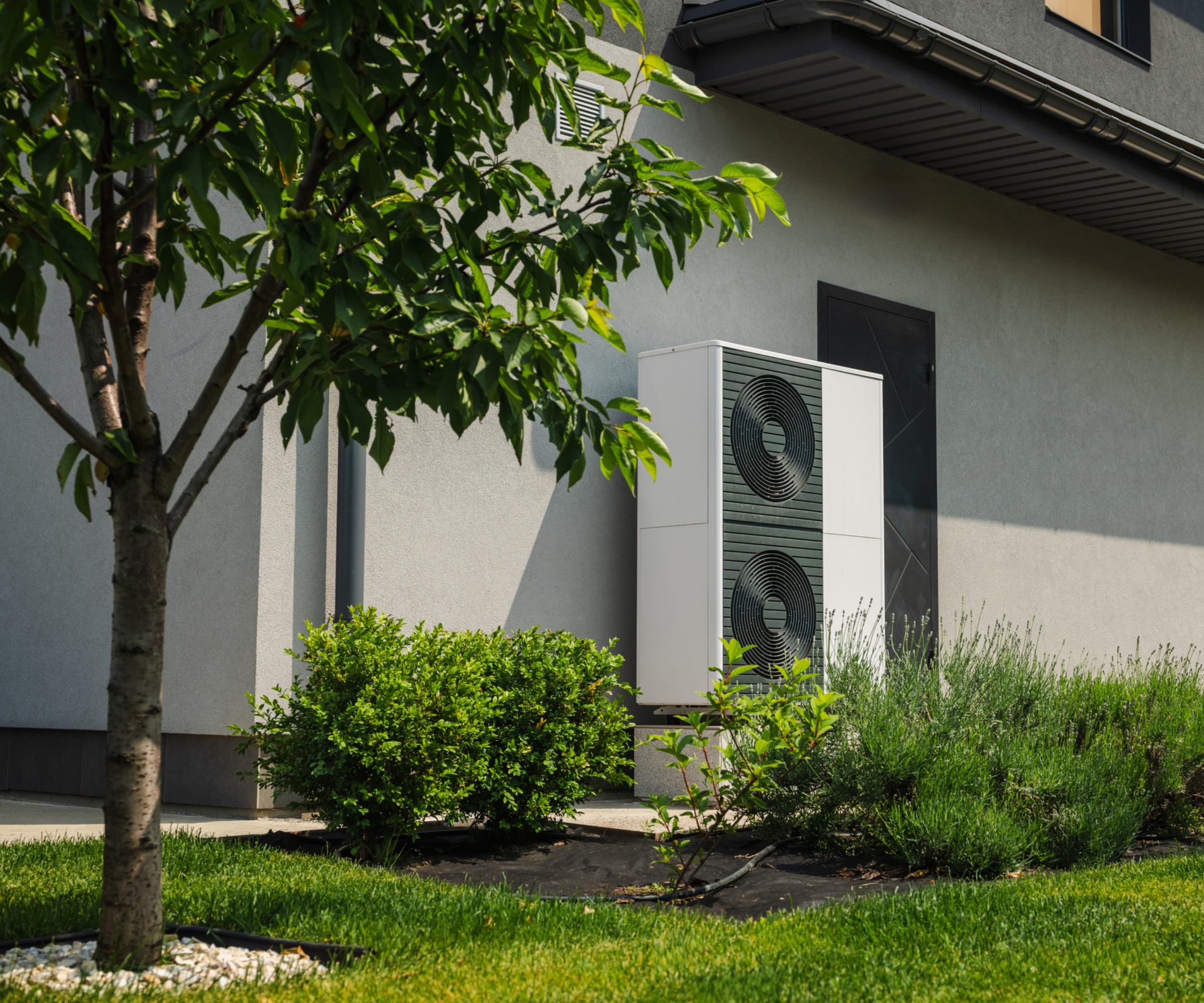 heat pump installed outside modern home with pale grey walls, trees and shrubs planted in lawned garden