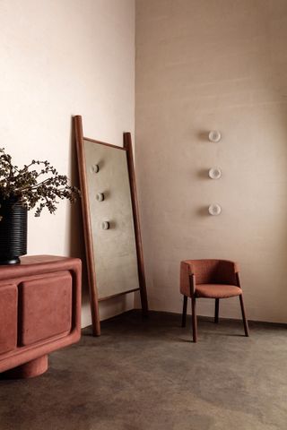The corner of a room with light mauve walls, grey carpet and furnitures (Brown wood framed mirror, Mauve velvet ottoma with a black vase with a plant. Mauve textiled round armed chair with 4 brown wood legs.