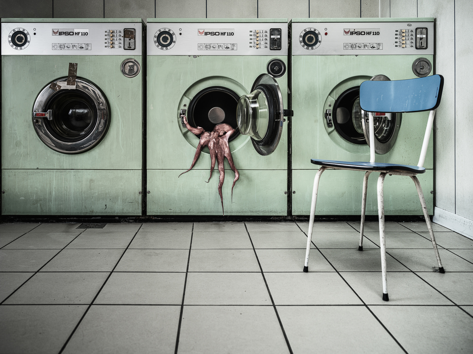 A green laundromat with three washing machines, one displaying an octopus, and a blue chair on tiled flooring