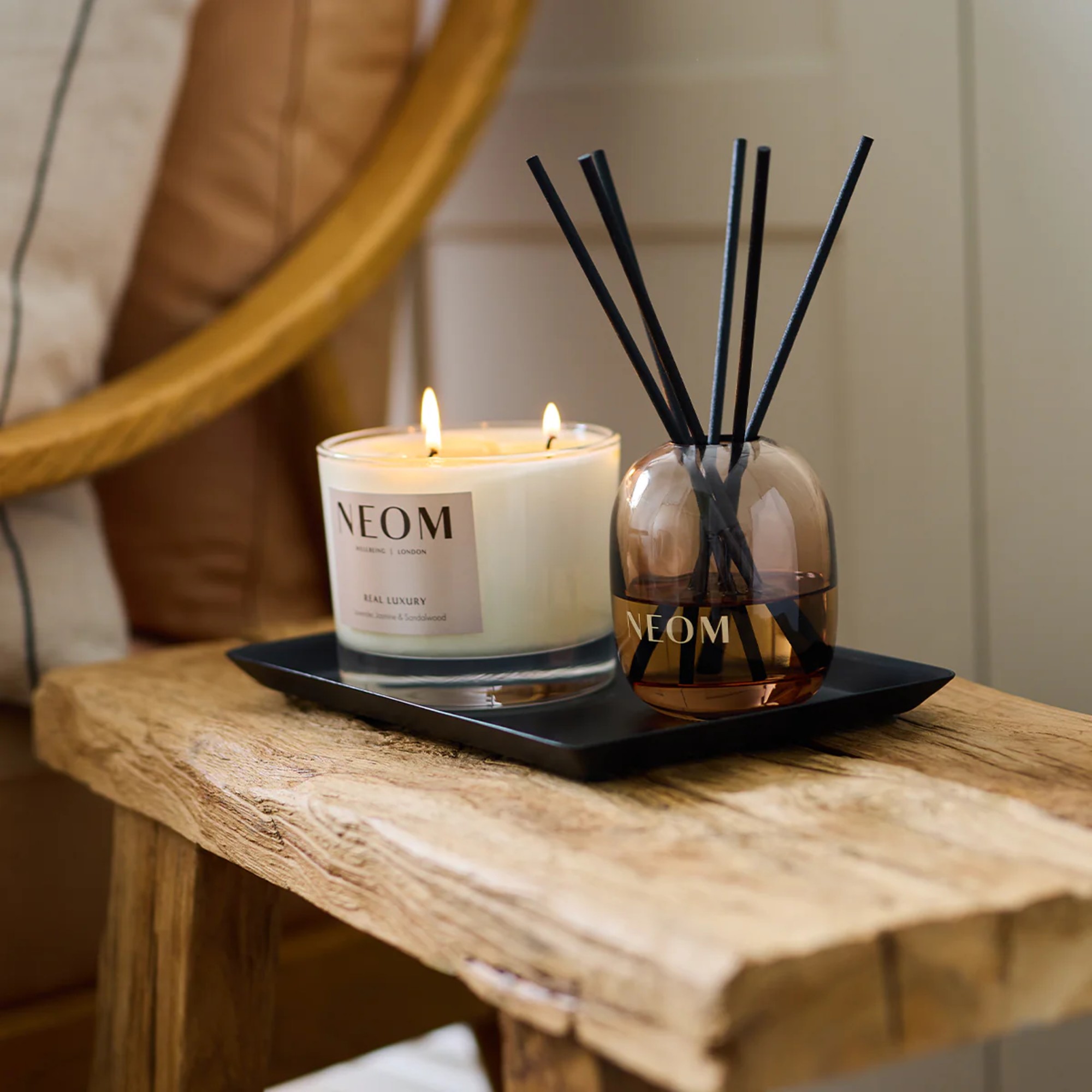 A three-wick Neom Wellbeing candle and reed diffuser on a rustic wooden side table